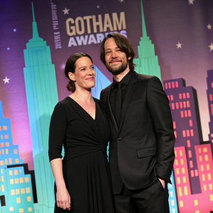 Franka Potente and Derek Richardson at the Gotham Awards. Franka in black dress, Derek in black suit. The backdrop features a colorful skyline.