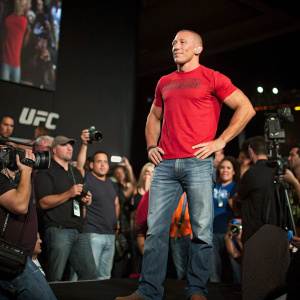 Georges St-Pierre, wearing a red t-shirt and jeans, at a UFC press conference. Crowd of fans and media in the background.