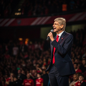 Arsene Wenger, dressed in a dark suit with a red tie, gives an impassioned speech. Crowd of fans in blurred background. Indoor arena