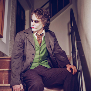 Concept art of the Joker from Batman series. Sitting on a dark, shadowy ledge. Pensive expression on his face. Wears a dark suit with a green vest and tie. His hair is styled unusually and face painted pale white. Background is dark and mysterious.