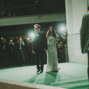 James Gunn and Adria Arjona at a movie premiere. He wears a black tuxedo, she in a long silver gown. Crowded red carpet with flashing cameras.