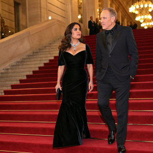 Salma Hayek, dressed in a figure-hugging black velvet gown, and François-Henri Pinault, in a sleek dark suit, walk the Met Gala red carpet. Grand staircase and golden chandeliers in the background