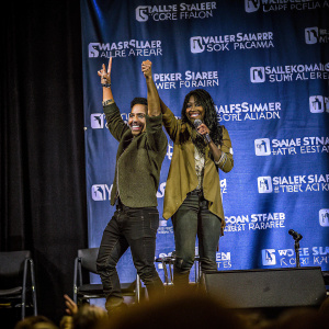 Ray Santiago and Jill Marie Jones hosting an event at Walker Stalker Con Atlanta. They stand on a stage, posing with a backdrop featuring the event's logo.