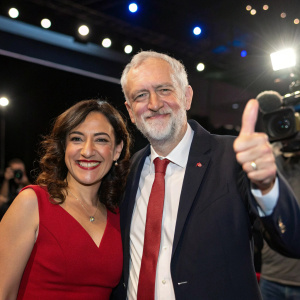 Jeremy Corbyn and Laura Alvarez, dressed formally, smiling and giving a thumbs-up at a press conference. Bright lights and cameras flashing in the background.