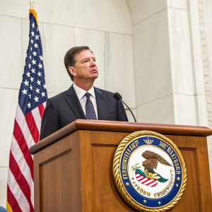 James Comey, in dark suit and tie, addressing a congressional hearing. Wooden podium with US flag and Department of Justice seal. White backdrop, serious expression