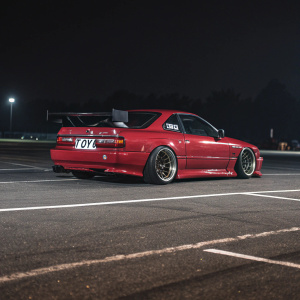 Custom Nissan Silvia S13 K drift car. Shiny red paintjob. Parked on empty asphalt lot with dark background. Widespread body, sponsor logos including Toyo tires, large rear wing.