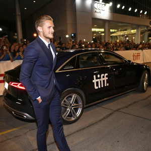 Alexander Ludwig, wearing a navy blue suit, at the Toronto International Film Festival. Standing beside a luxurious Audi car. The background is filled with flashing cameras and excited fans.