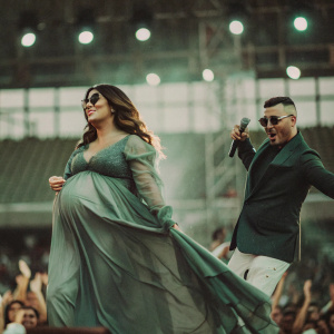Wisin and Yomaira Ortiz Feliciano, on the stage, she's pregnant and in a flowing green dress, he's in a dark suit and shades. Flashing lights, fans screaming, excitement
