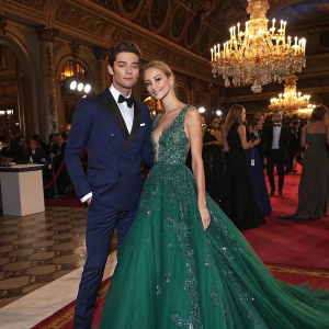 Francisco Lachowski and Jessiann Gravel Beland at a glamorous gala. Francisco in a tailored navy suit. Jessiann in a floor-length emerald green gown with intricate beadwork. Lavish ballroom setting with chandeliers and guests.