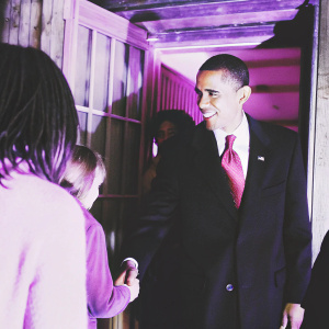 Barack Obama, former US President, at a fundraising event. Black suit, red tie. Greeting attendees. Dimly lit room.