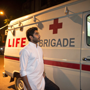 Abhishek Bachchan standing outside an ambulance during a charity event. Ambulance has a red cross symbol and 'Life Brigade' written in bold letters. Indian actor, wearing a white shirt, with a serious expression. Dark-skinned, medium build.