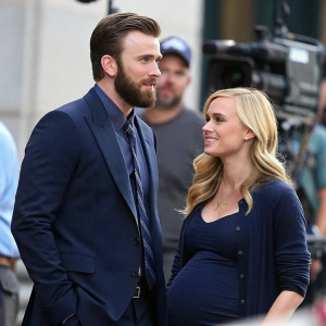 Chris Evans, bearded and in a dark blue suit, stands beside a pregnant woman with blonde hair. They are on a movie set. Blurred crew members in background. Candid shot.