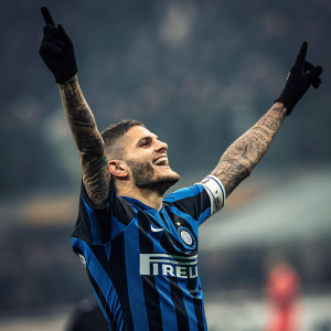 Mauro Icardi, celebrating a goal with arms raised and big smile. Blue and black striped jersey. Background blurred stadium spectators.