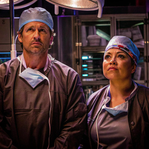 Patrick Dempsey and Sara Ramirez in surgical attire, standing in front of a sterile operating room. Both looking serious with hospital equipment visible in the background.