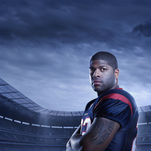 Arian Foster, African American football player, intense gaze, cropped hairstyle, tattoos on arms. Wearing Houston Texans jersey. Indoor stadium, spectators visible.