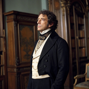 Carlo Puri as Viscount Andrea Cavalcanti from 'The Count of Monte-Cristo', standing solemnly. White man with curly hair. Formal attire: black jacket, white shirt, fancy necktie. Set design includes large antique wooden furniture.