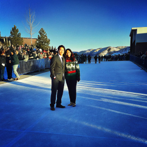 Randall Park and Jae Suh Park attending the Sundance Film Festival premiere. Randall in a classic dark suit, Jae Suh in a vibrant Christmas-themed sweater. Crowd and photographers in the background.