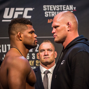 Alistair Overeem and Stefan Struve face off in intense pre-fight moment at UFC event. Promotional banner and referee in background, professional sports photograph.