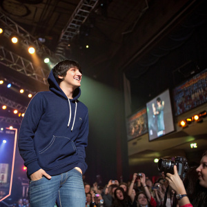 Mitchel Musso, at a concert venue, dressed in a navy blue hoodie and jeans. Surrounded by screaming fans and flashing cameras.