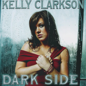 Promotional shot for Kelly Clarkson's 'DARK SIDE'. Kelly, serious expression, medium brown hair, red off-shoulder top, adjusting her attire with intense focus. Moody and dramatic backdrop, with album text 'KELLY CLARKSON' and 'DARK SIDE' in bold.