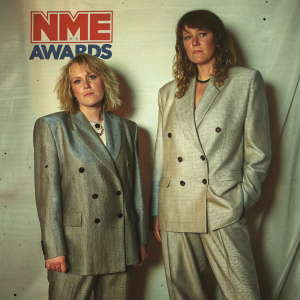 Edith Bowman and Jo Wiley at the NME Awards, both wearing tailored suits. The background features the NME Awards logo against a plain white backdrop.