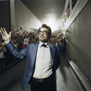 Robert Downey Jr. at Comic-Con in San Diego. Blue suit, glasses, and black belt. Crowded venue with fans.