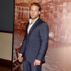 Jai Courtney in a navy blue suit, at Venice Film Festival. Behind him is a blurred cityscape