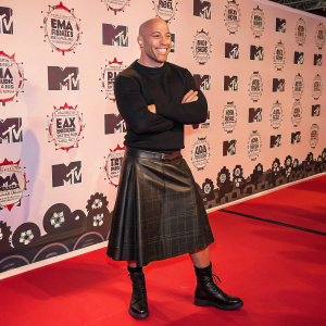 Vin Diesel, Caucasian, bald, at MTV Europe Music Awards. Dressed in black sweater, leather kilt. Confident pose, broad smile. Red carpet with repeating logos and monkey illustrations backdrop.
