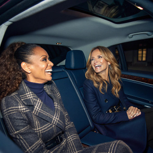 Mel B and Geri Halliwell inside a luxury car, laughing and chatting. Dark blue seats and windows tinted