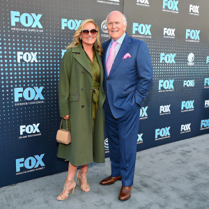 Tammy Bradshaw and Terry Bradshaw at the annual Fox Upfront event. She's in an olive-green coat with sunglasses and beige accessories. He's in a blue suit with a pink-toned tie and soft-pink pocket square. Standing on gray carpet with backdrop of repeated logos and digital pattern.