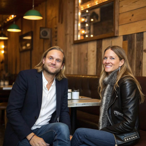 Chad Muska and Vanessa Traina at a cozy cafe. Casual outfits, friendly expressions. Chad in a dark blue blazer and white shirt, Vanessa in a black leather jacket and a faux fur scarf. Background is rustic wooden walls and warm lighting.