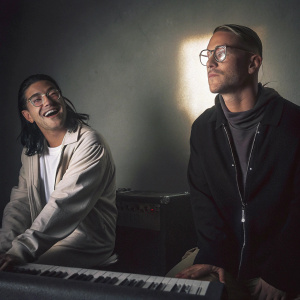 Skylar van der Waal and Thomas Wesley Pentz, also known as Diplo, are seen in a promotional photo shoot. Skylar, known by his stage name Skrillex, sports a casual outfit and a light-hearted expression. Thomas, on the other hand, dons a more serious demeanor in his dark ensemble.