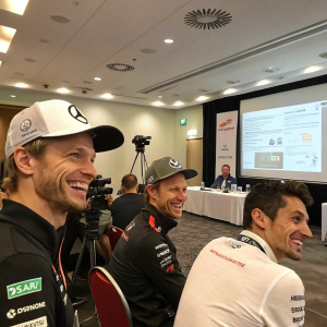 Formula 1 drivers Nico Hulkenberg, Jenson Button, and Esteban Ocon at a motorsports conference. Conference room with projector screen and charts on the wall. Smiling for photographers