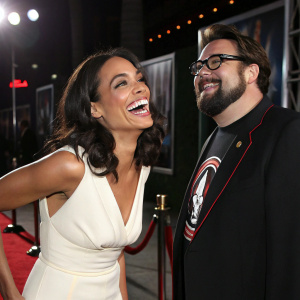 Rosario Dawson, Hispanic actress in a white dress, laughing. Kevin Smith, Caucasian man with beard and glasses, smiling. They are at a movie premiere with dramatic stage lighting and velvet ropes in the background.