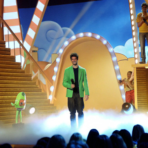 Grant Gustin, wearing a green blazer and black t-shirt, accepting an award. The stage is filled with animated characters and bright lights from the Nickelodeon Kids' Choice Awards.