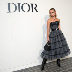 Amelia Windsor, model, wearing a black and gray Dior dress with a ruffled skirt, standing in front of a stark white wall with the Dior logo prominently displayed. She poses elegantly with a belt and black boots.