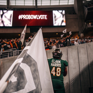 Clark Harris, clad in a vibrant green jersey with number 48, stands before a crowd at a stadium. The backdrop is a sea of fans holding up banners and waving flags. The hashtag #ProBowlVote lights up on large screens.