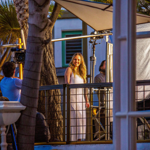 AJ Michalka in a candid moment. She has long, wavy blonde hair, a friendly smile. Wearing a simple white dress. On-set between takes at an outdoor filming location.
