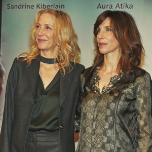 Photocall event featuring Sandrine Kiberlain and Aura Atika. Both women in dark outfits. Sandrine with blonde wavy hair, black choker. Aura with warm-toned complexion, brown hair. White background with text elements.