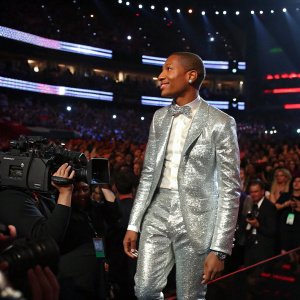 Pharrell Williams at the Grammy Awards, dressed in a silver sequined suit. Large crowd and flashing cameras in the background.