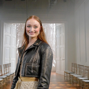 Sophie Turner, smiling warmly in a black leather jacket, against a minimalist white backdrop. Fashion show for Dior and EuroPAC collaboration.