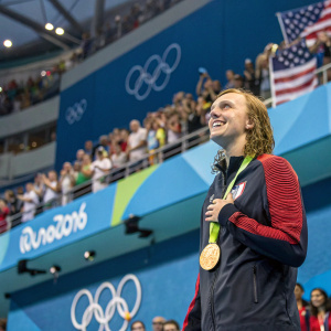 Katie Ledecky, wet hair cascading down her shoulders, standing in the Olympic podium amidst a crowd of cheering spectators.