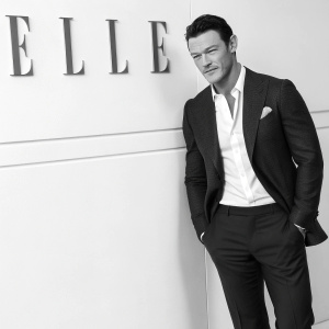 Luke Evans, a well-dressed gentleman, standing in a stylish black and white photograph. He is in front of a wall adorned with the Elle logo. Professional photo shoot