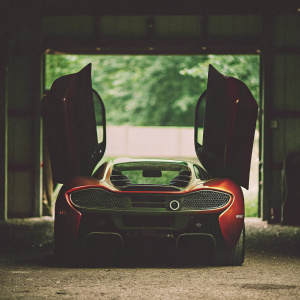 In a dimly lit garage, a sleek, crimson-hued McLaren unfolds its rear wings, casting eerie shadows. Angular LED lights pulse with a menacing glow. The vehicle's honeycomb-patterned underbelly seems to whisper secrets of the past. Aerodynamic lines slicing through the darkness, the car's precise curves glisten under the soft lighting, showcasing the futuristic masterpiece in haunting detail.