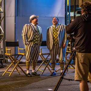 Channing Tatum and Jonah Hill in comedic outfits. Behind-the-scenes of movie set. Directors' chairs, stage, plain backdrop. They're laughing, relaxed pose, microphone present.