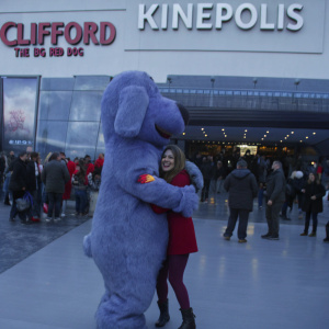 Beatriz Jarrin, wearing a red outfit, enthusiastically hugging a giant purple mascot. Crowded event, movie premiere for 'Clifford: The Big Red Dog' at Kinepolis.