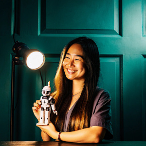 Young Asian woman, with long brown hair, posing with a Frieza figurine. She's smiling warmly in a dark room with only a spotlight illuminating her.