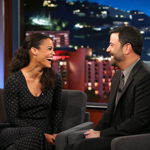 Paula Patton and Jimmy Kimmel having a lively discussion. Studio setting with warm cityscape backdrop. Soft, focused lighting