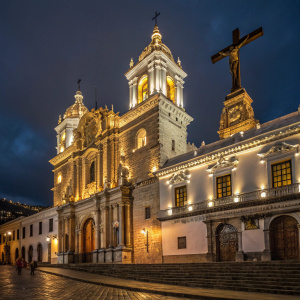 In the heart of Quito, a majestic building stands out. It features dazzling stonework, illuminated by golden lights. Intricate carvings and statues adorn its facade. A large crucifix is prominently displayed at one corner.