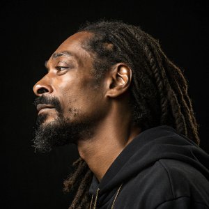 Close-up portrait of Snoop Dogg with prominent dreadlocks and beard. Solemn expression against stark black backdrop.